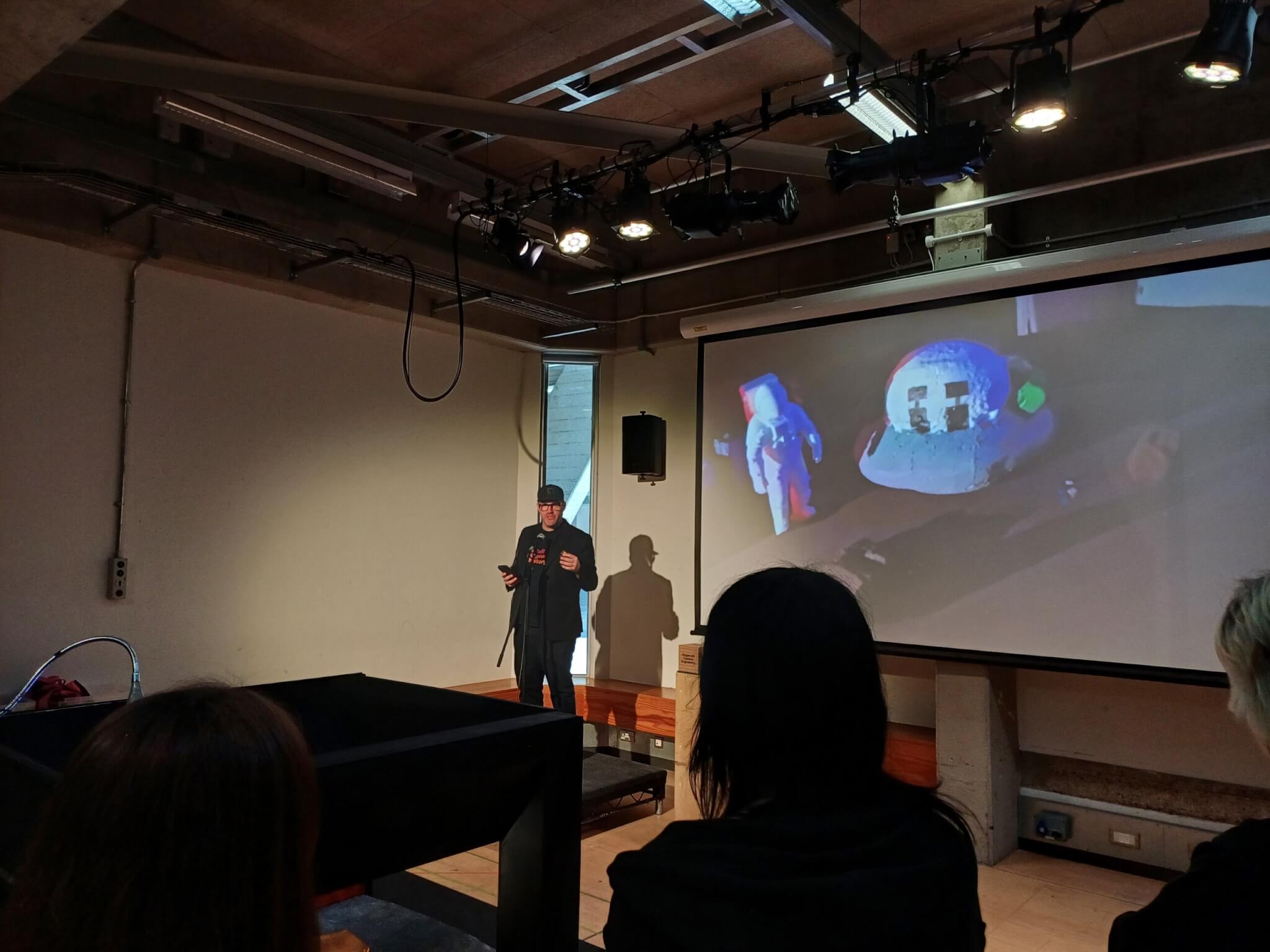 Chris Mitchell, ESA Principal, in a black baseball cap and clothes, on a small stage speaking to the audience. Behind him is a projector showing an alien ship and astronaut -- one of the staging designs.