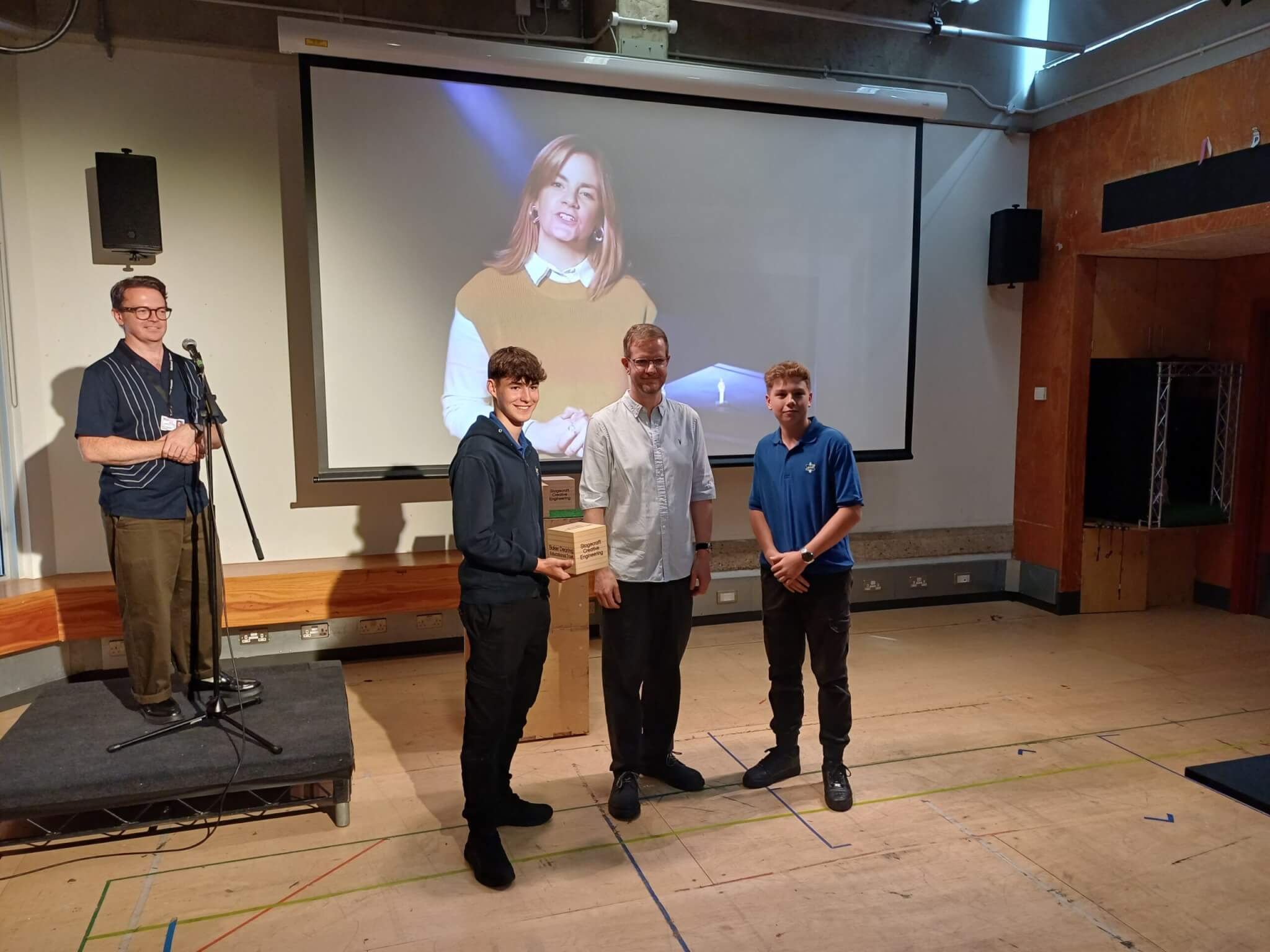 Two UTC Warrington students in branded apparel stood in a row for photos with Alex Eales before a projector screen.