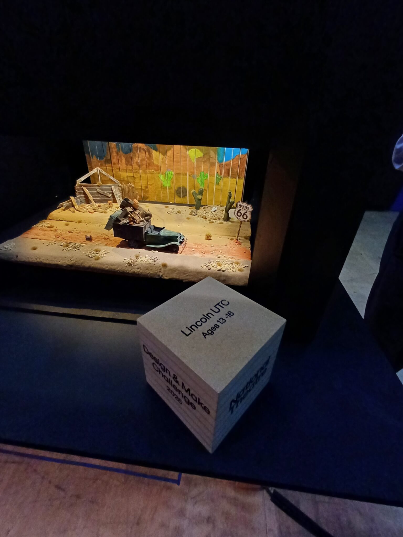 A wooden cube. Text on its top reads 'Lincoln UTC Ages 13-16'. Text on its left face reads 'Design and Make Challenge 2025'. 'National Theatre' is inscribed on its right face. Behind the cube award is the students' design, consisting of a Jeep in a desert background, with a Route 66 sign ahead of the vehicle.