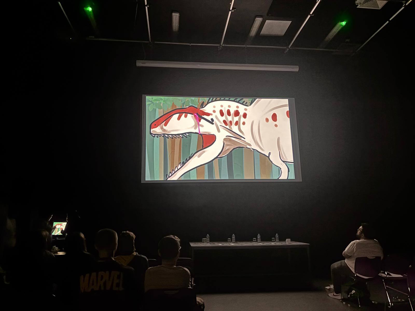 Students sat in darkness watching a projector screen, showing an animation of a beige dinosaur-like creature with red spots that has been pierced with a spear and is crying out in pain.