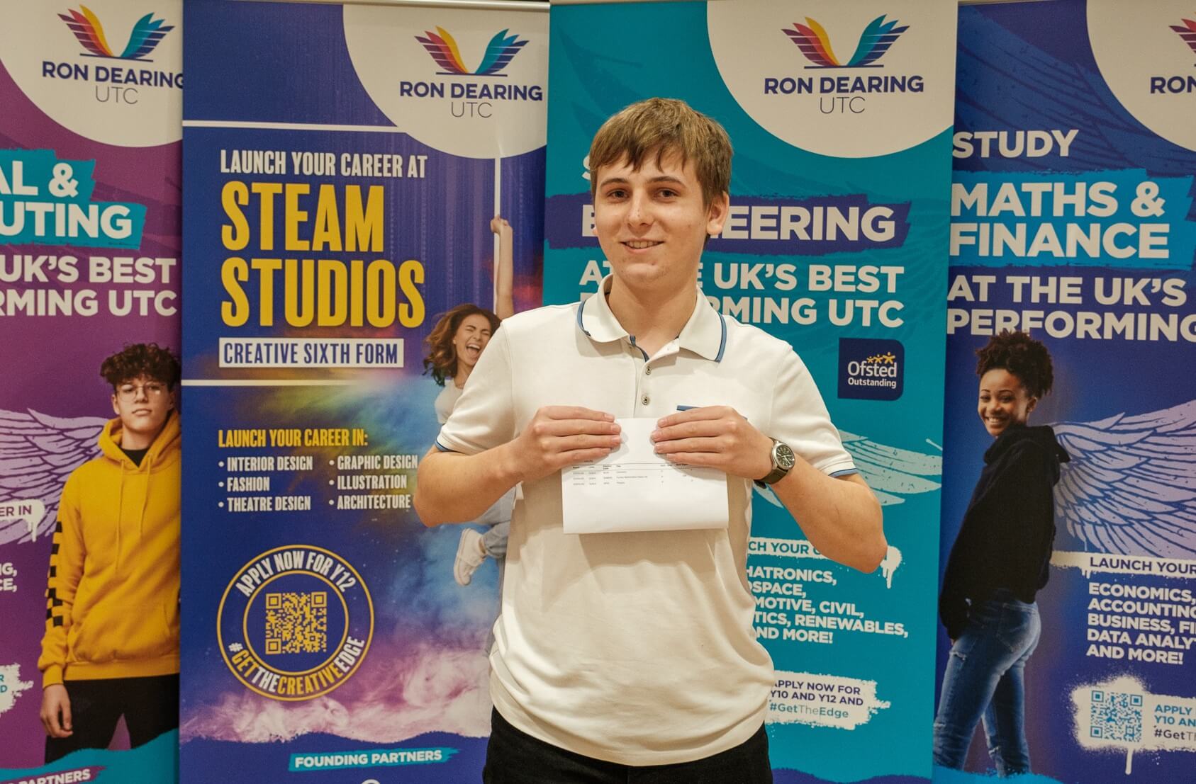 A young man proudly presents his results on a piece of paper in front of a selection of Ron Dearing UTC branded roller banners. He looks confident and enthusiastic