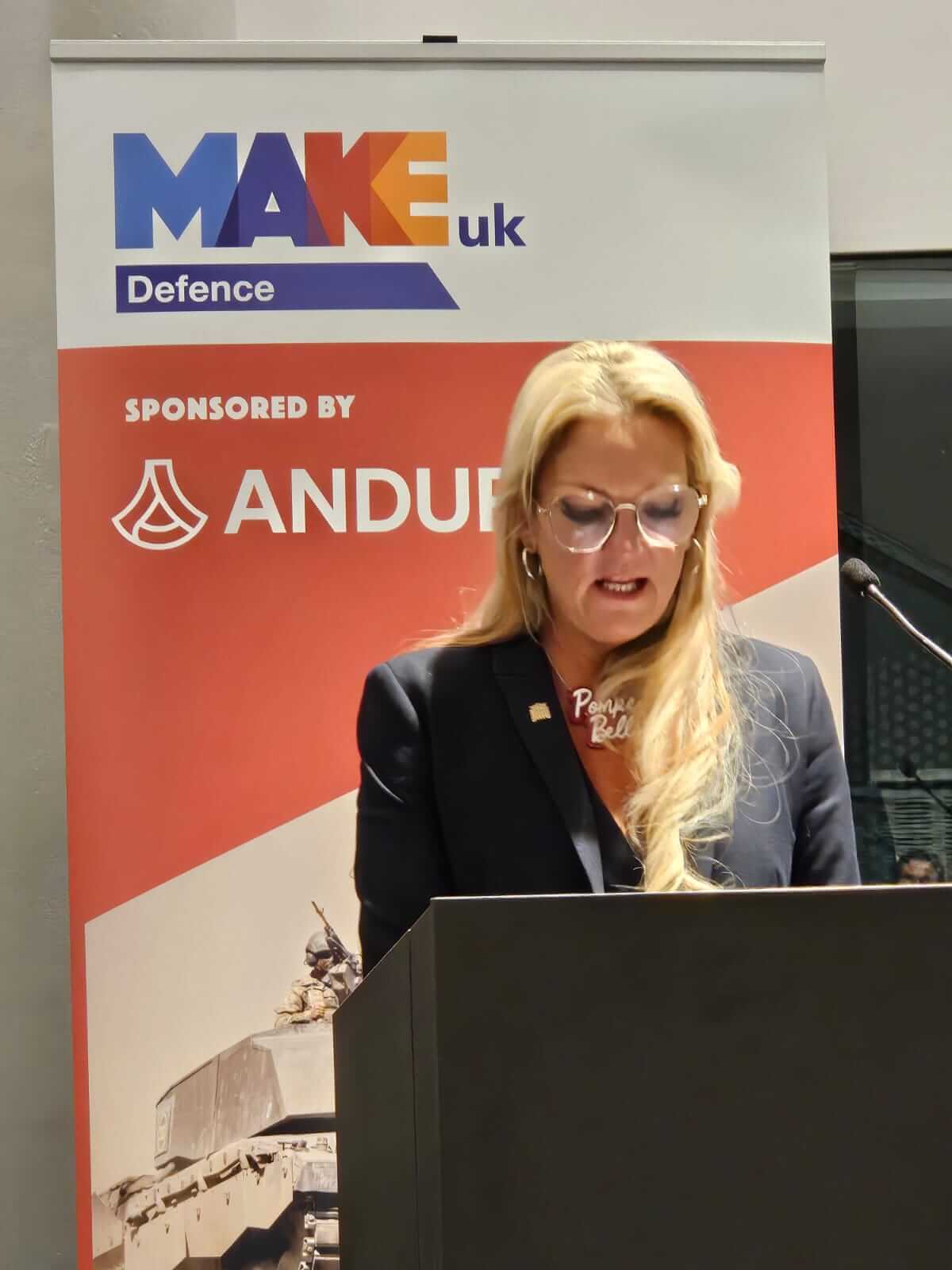 A woman stood speaking at a podium before a Make UK Defence banner.