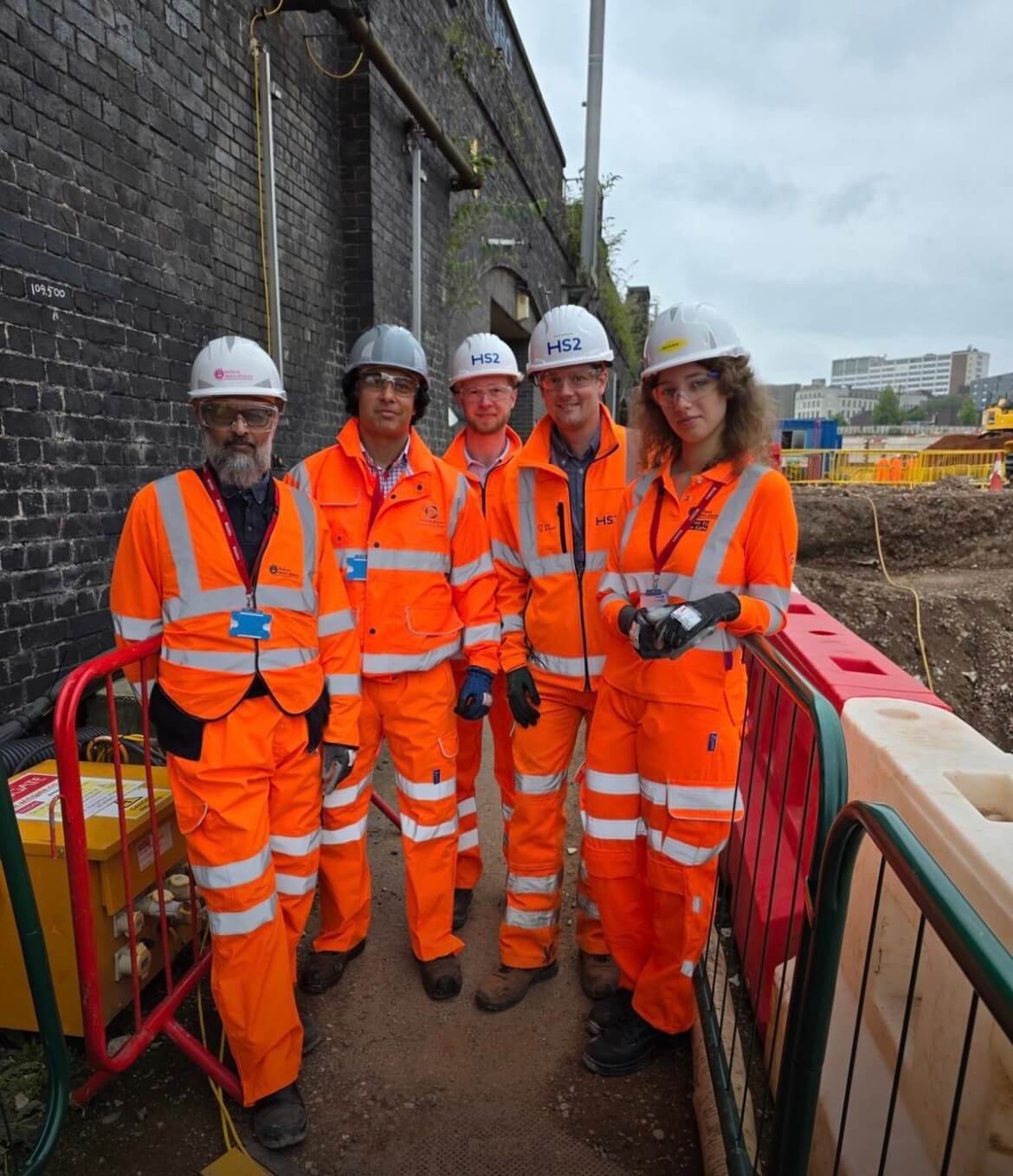 A team dressed in hi-vis orange work uniforms lined up alongside a fence with a brick wall in the background.