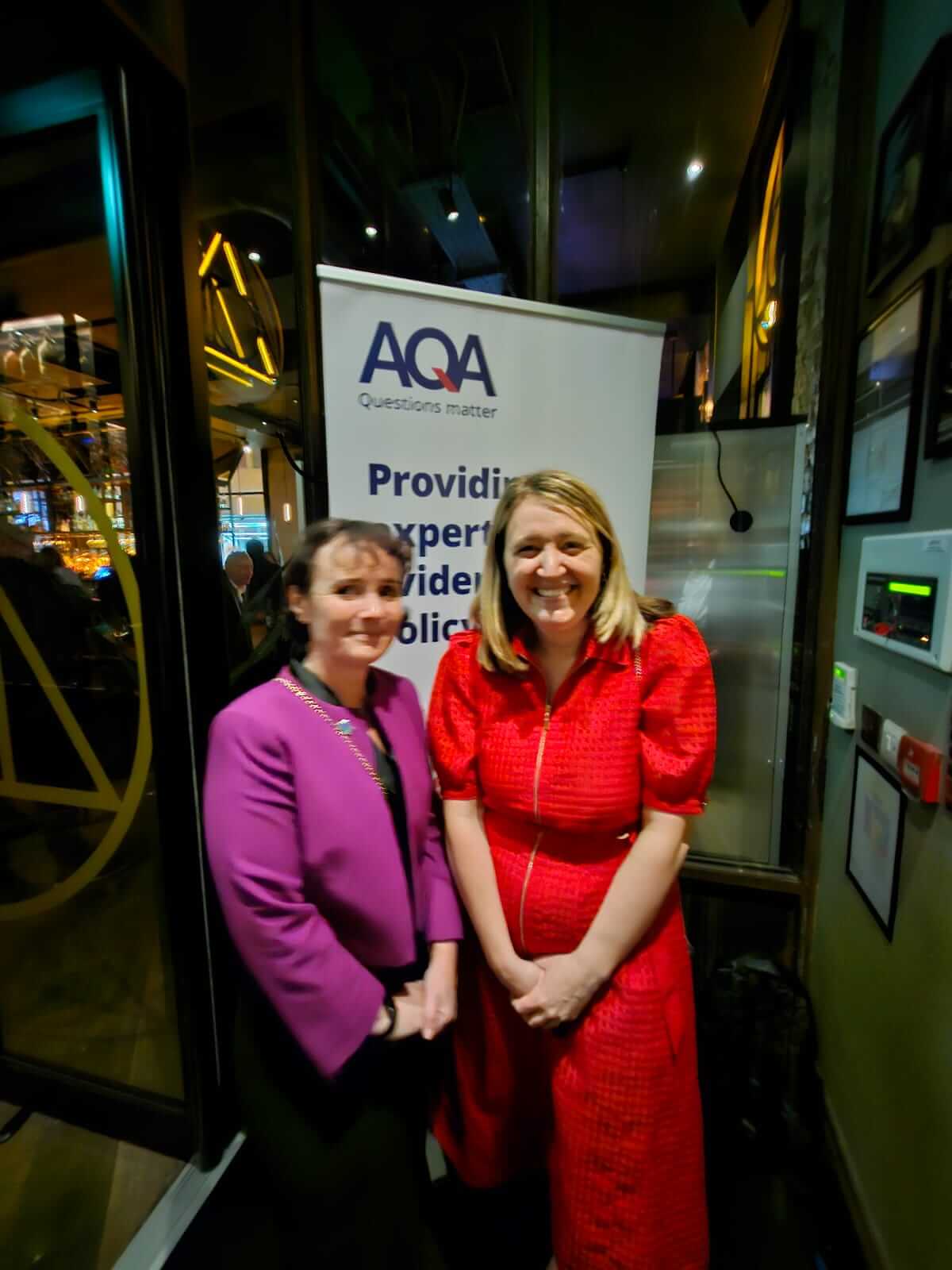 Two women stood before an AQA banner in the foyer outside a bar area.