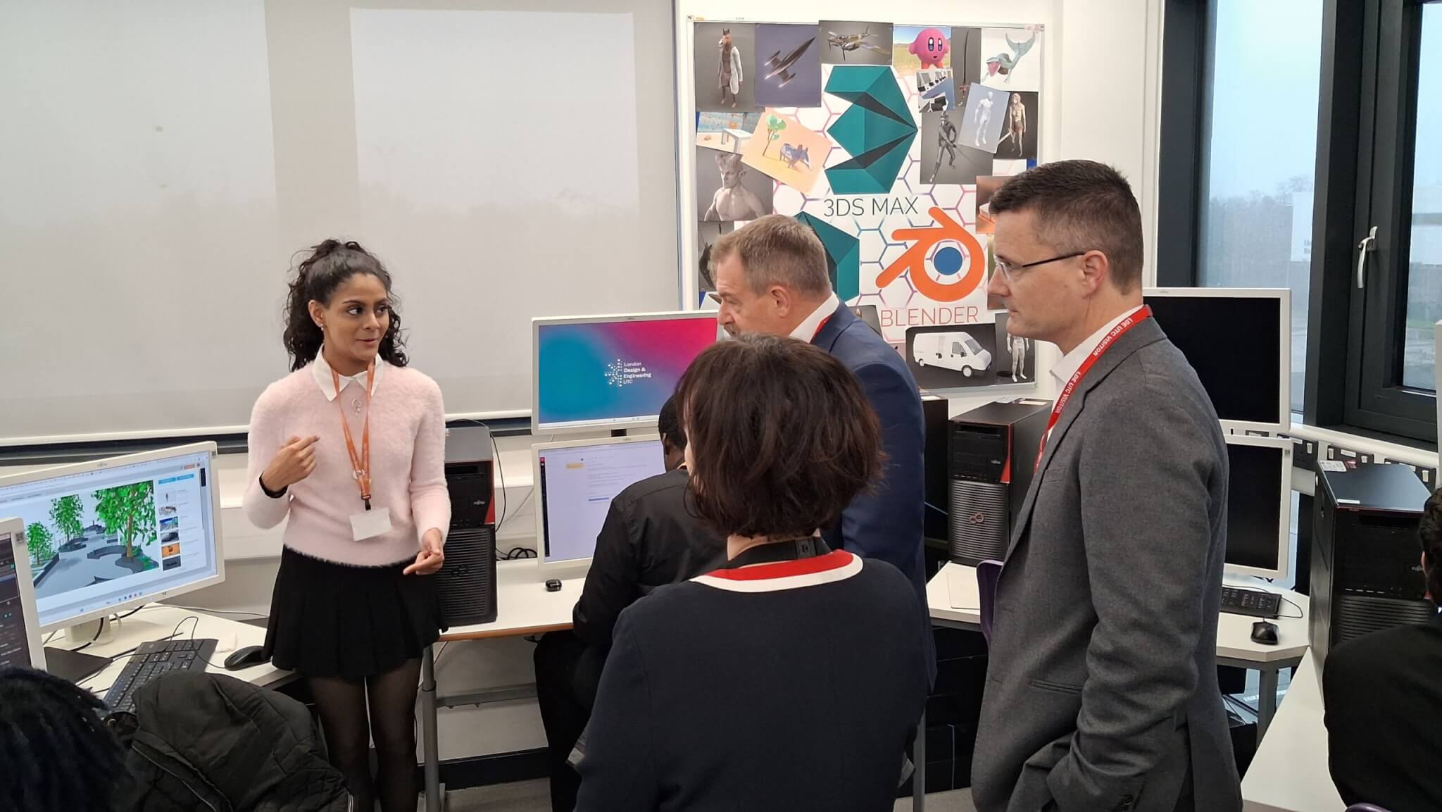 A female student in a pink jumper presenting to two men in suits and a woman, amidst a busy classroom environment.