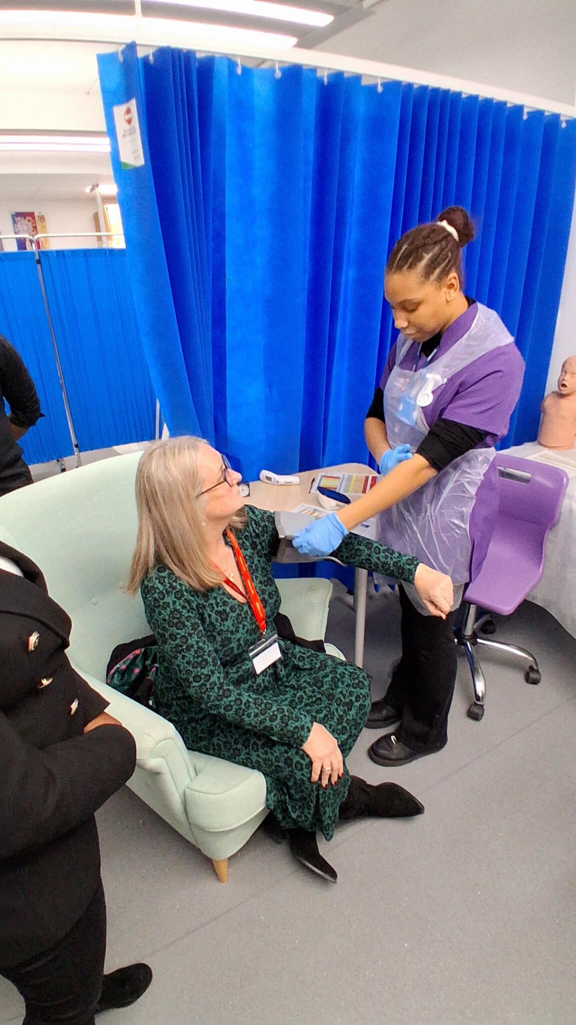 A woman sat in a green armchair with a blood pressure test band around her arm and a female student stood over her.