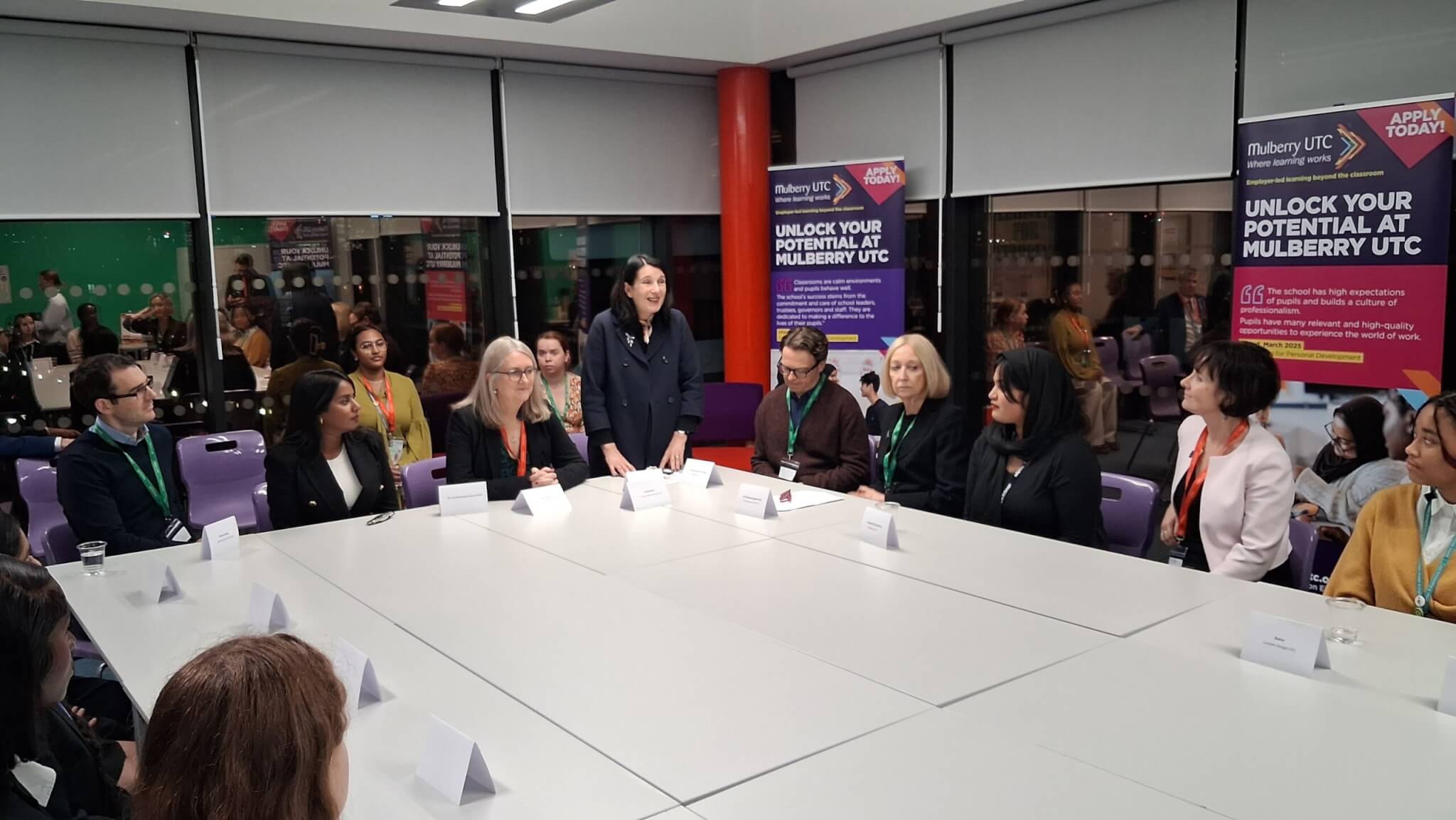 A group of women sat around a large table with one woman stood the furthest away from the camera giving introductions.