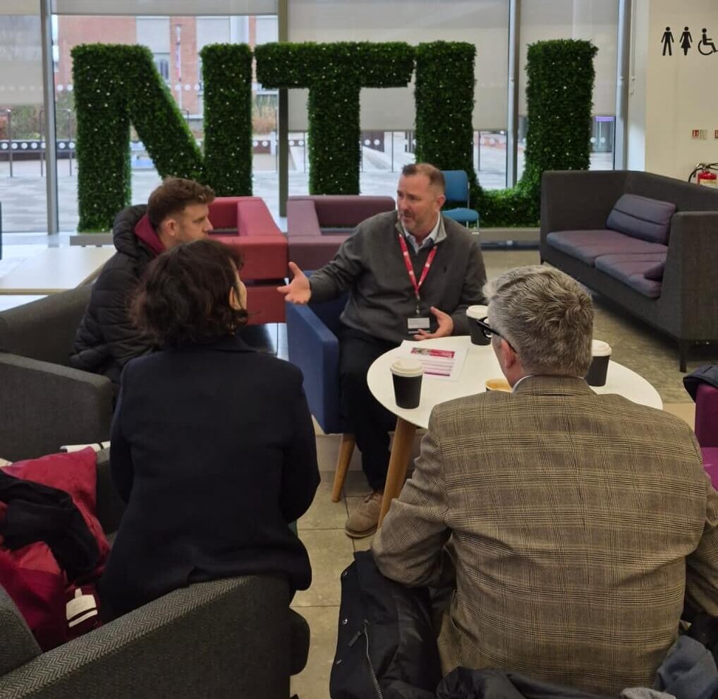 A group of people seated in chairs in an atrium, with a sign made of turf displaying "NTU" in the background.