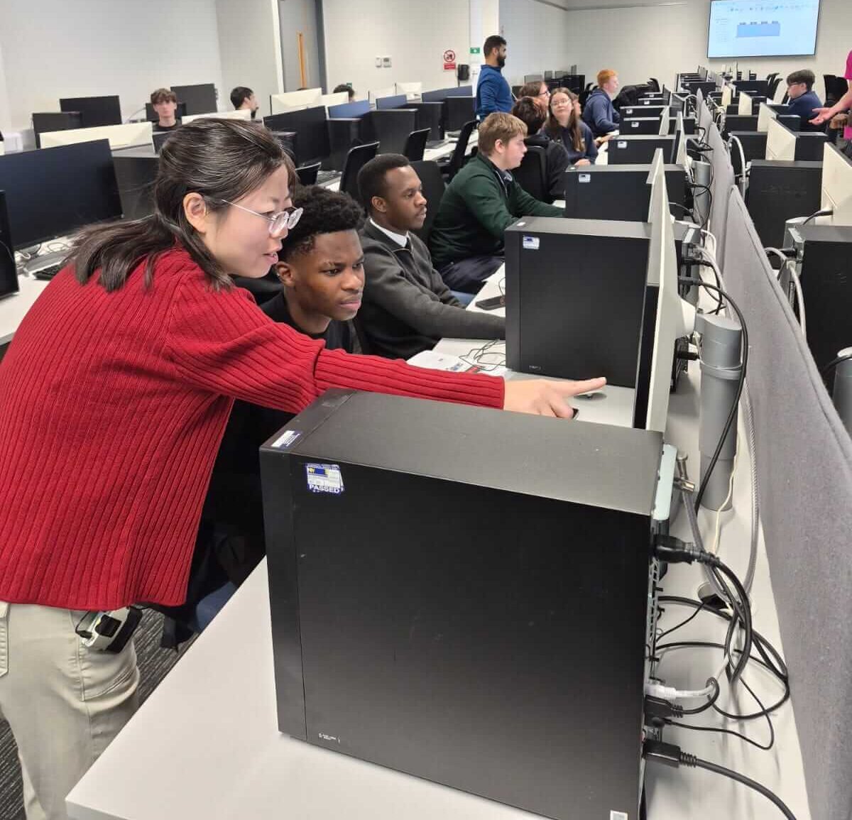 A lecturer stood over a young student, pointing at something on the student's computer screen. Students are sat before a row of computers in the background.