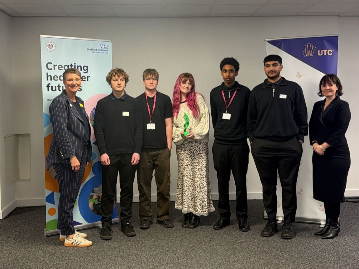 Another group of students. The centre student is holding a green trophy. Brown is stood on the left, before the NHS banner, and Ambrosi on the right, by the UTC banner.