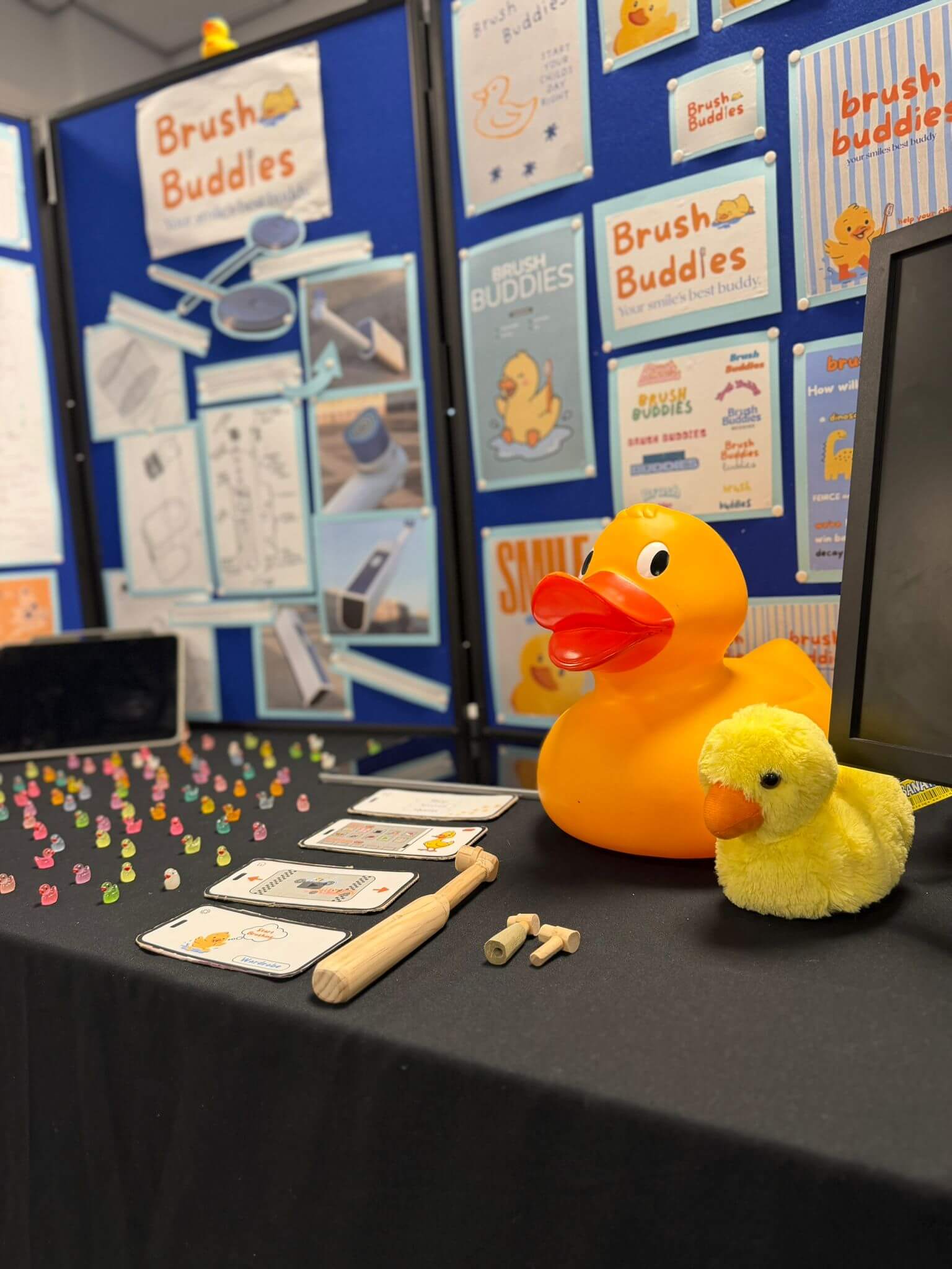 A table with a large rubber duck atop next to a smaller, fluffier duck. There is also a wooden tooth brush and toothbrush heads, a range of small, multi-coloured items, and a large display board in the rear with the legend 'Brush Buddies.'