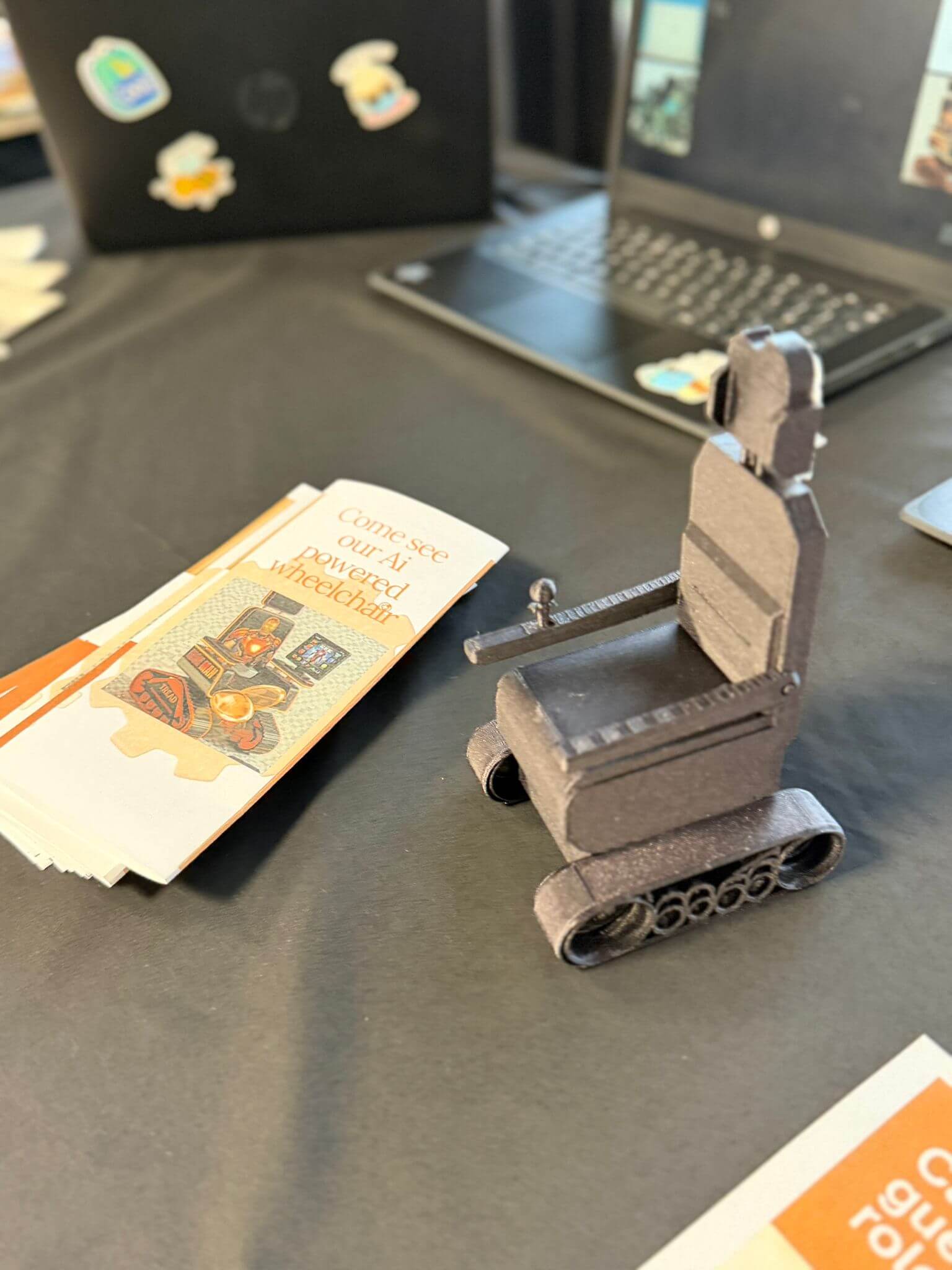 A tiny model wheelchair sits on a table next to an open laptop and a pile of leaflets titled 'come and see our AI powered wheelchair'