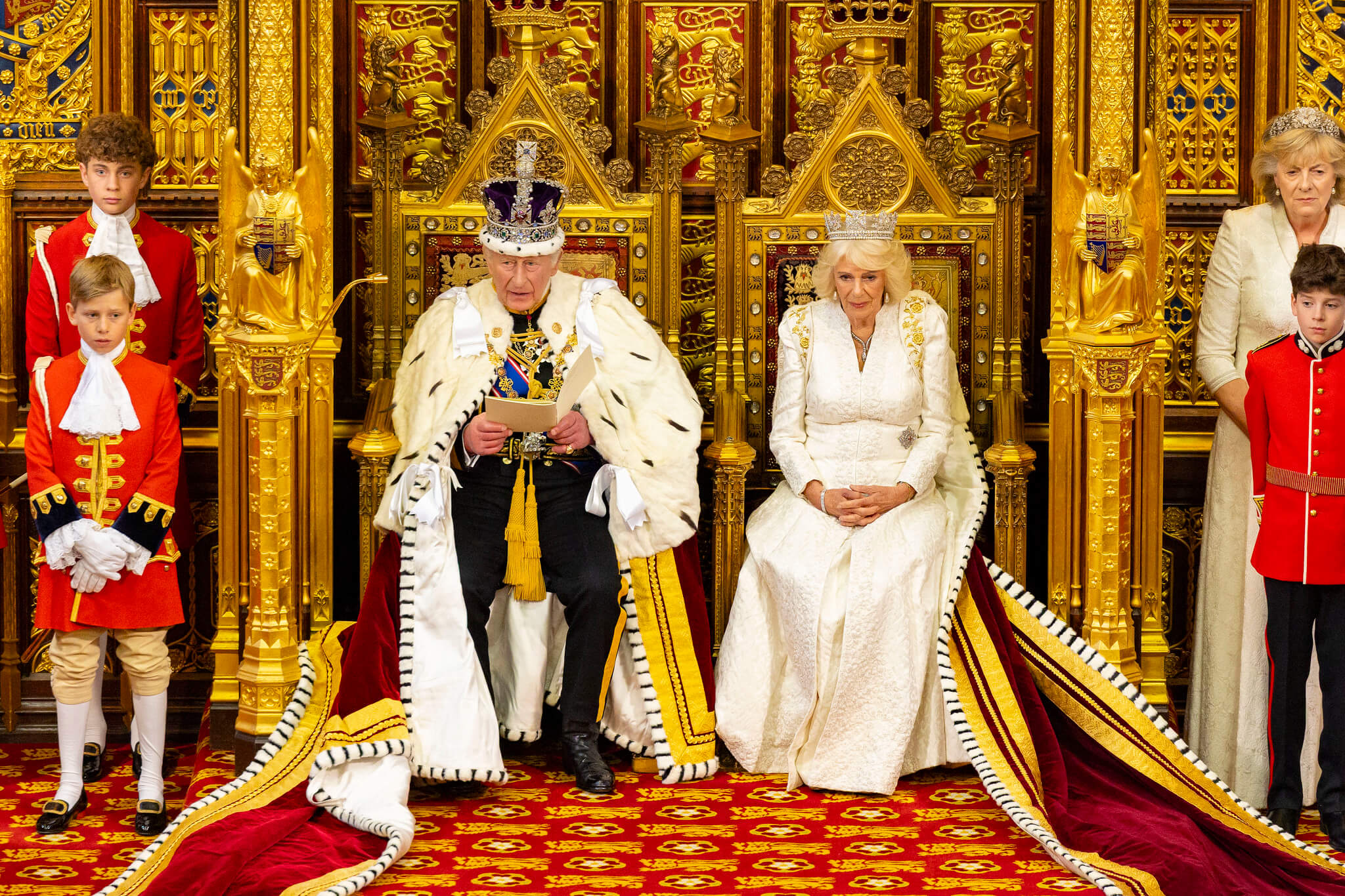 ©House of Commons King Charles and Queen Camilla sit on thrones against gold decoration. Pages and ladies in waiting flank them.