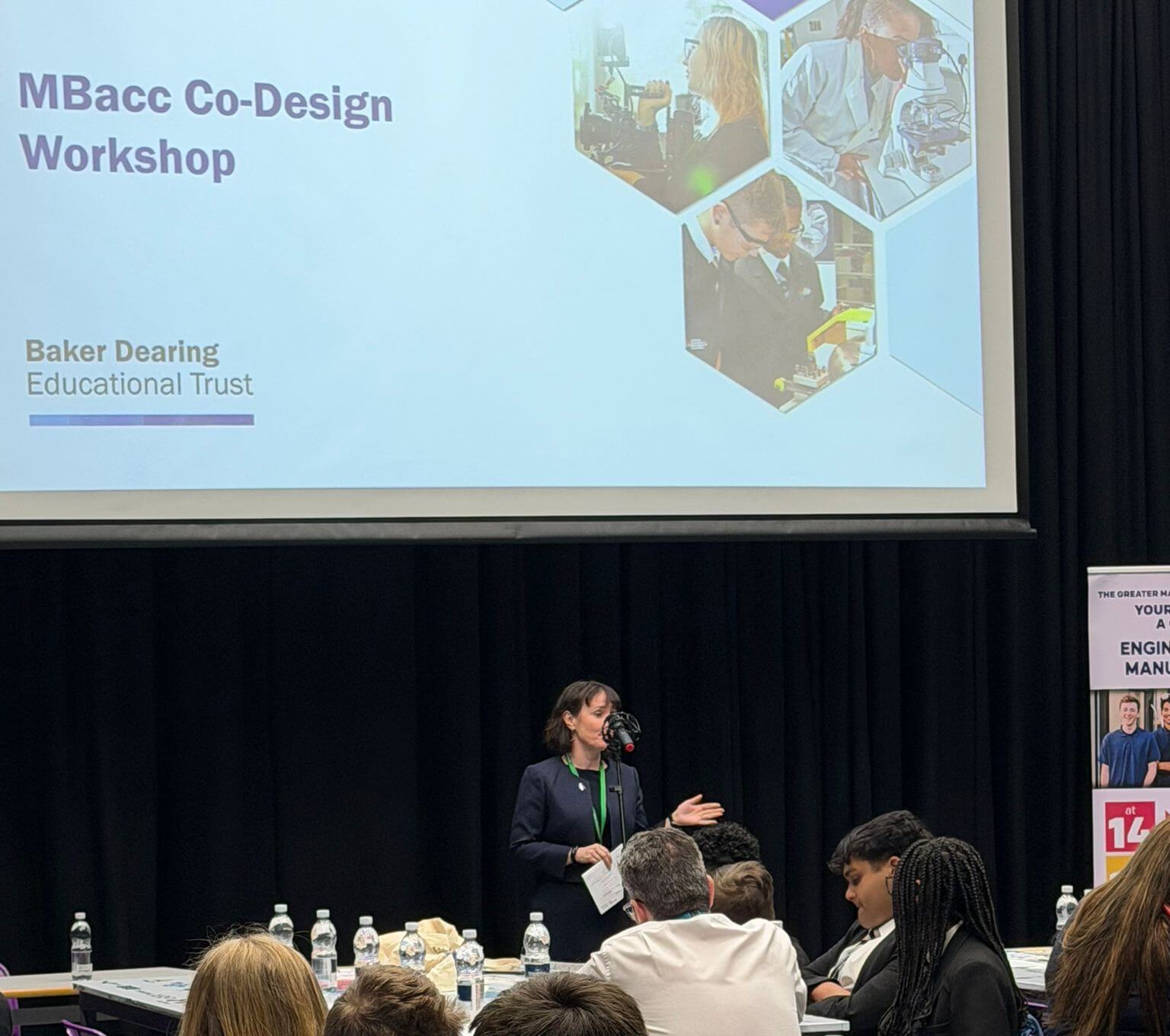 A woman addressing a room of people. Above her is a projector screen reading 'MBacc Co-Design Workshop' with the Baker Dearing logo.