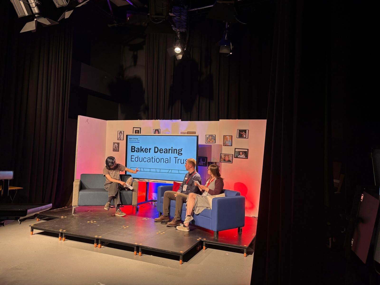Three people on a set, sat on sofas with the interviewer on the left-hand sofa and two individuals on the right-hand sofa. A TV sits between them showing the Baker Dearing logo. A black curtain hangs around the set.