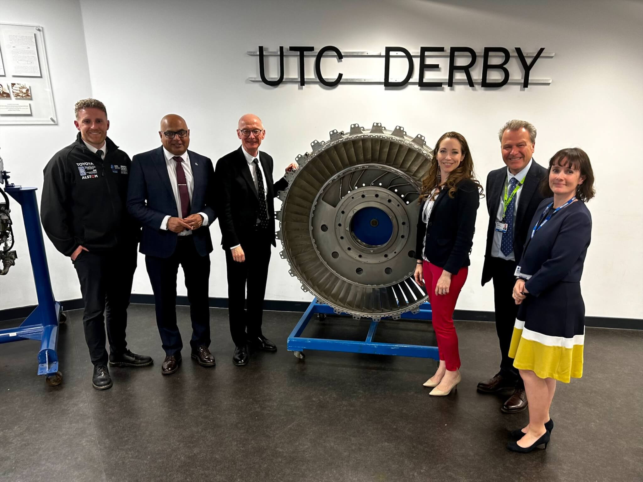 A group of adults gathered around a large gear, before a sign reading 'UTC Derby'. L-R: Lee Kirkwood, Baggy Shanker, Pat McFadden, Catherine Atkinson, Nick Crew, Kate Ambrosi.